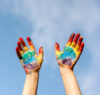 two hands coated in rainbow paint held against a blue sky